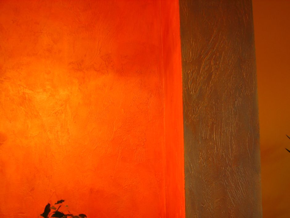 Fragment pomarańczowej ściany z wyraźną fakturą obok beżowego filaru z dekoracyjną strukturą. W rogu widoczny fragment rośliny.